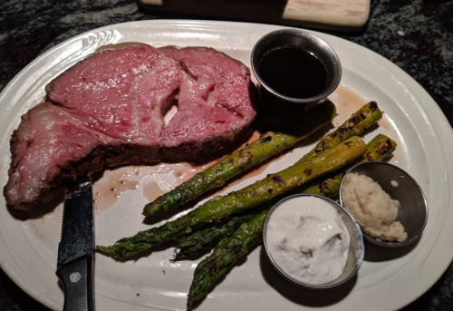 A plate with a juicy prime rib, grilled asparagus, and two dipping sauces on the side.