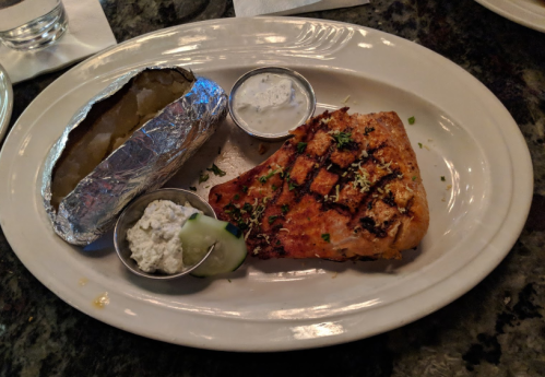 Grilled salmon fillet with herbs, served with a baked potato and dipping sauce on a white plate.