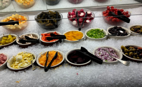 A variety of colorful toppings in bowls on ice, including fruits, vegetables, and cheeses, with serving utensils.