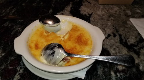 A bowl of crème brûlée topped with melting ice cream, served with two spoons on a marble countertop.