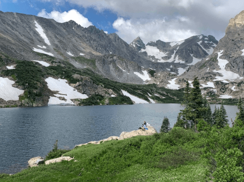 A serene mountain lake surrounded by snow-capped peaks and lush greenery, with two people sitting on a rock by the water.