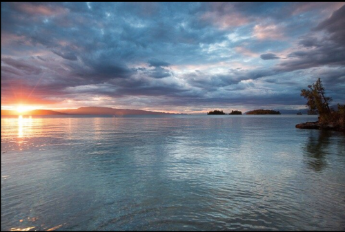 A serene lake at sunset, with colorful clouds reflecting on the water and distant islands on the horizon.