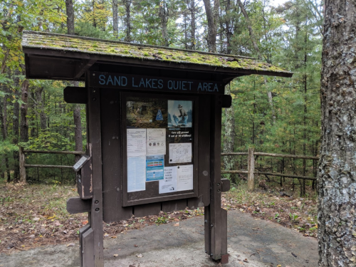 Information sign for Sand Lakes Quiet Area, surrounded by trees in a forested setting.