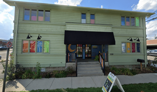 A green two-story building with colorful windows and a black awning, surrounded by greenery and a sidewalk.