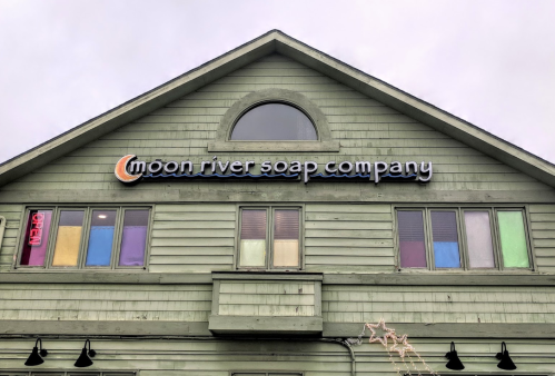 Sign for Moon River Soap Company on a green building with colorful windows, under a cloudy sky.