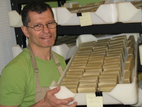 A man in an apron smiles while holding a tray of neatly arranged soap bars in a workshop.