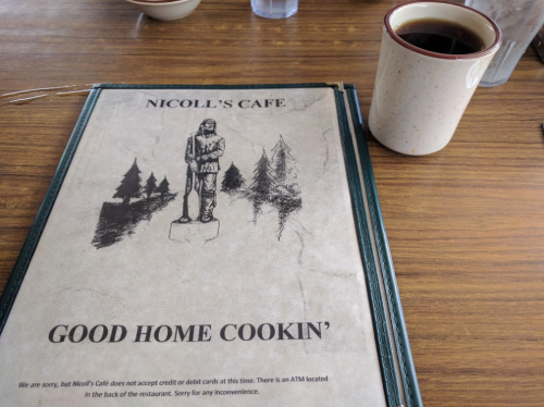A menu from Nicoll's Cafe featuring "Good Home Cookin'" and a cup of coffee on a wooden table.