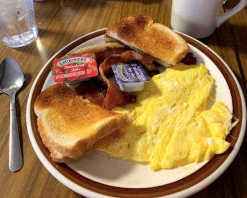 A plate with two slices of toast, bacon, scrambled eggs, and condiments on a wooden table.