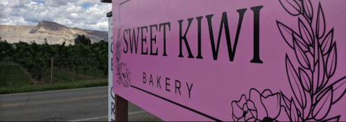 A pink sign for "Sweet Kiwi Bakery" with a scenic mountain and vineyard backdrop.