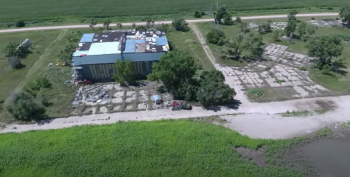 Aerial view of a large building near a road, surrounded by green fields and a river, with organized plots nearby.