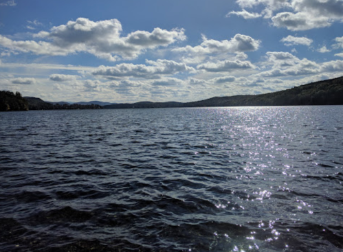 A serene lake under a blue sky with fluffy clouds, reflecting sunlight on the water's surface.