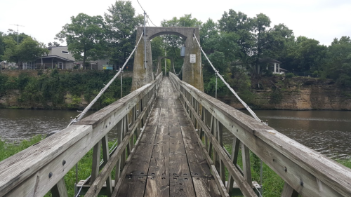 A wooden suspension bridge spans a river, surrounded by greenery and a rocky bank, leading to a tree-lined area.