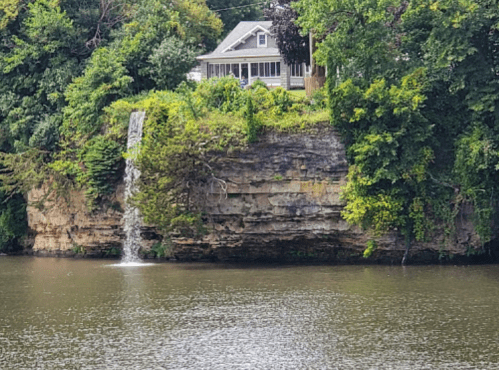 A waterfall cascades down a rocky cliff beside a calm river, with a house nestled among lush greenery above.