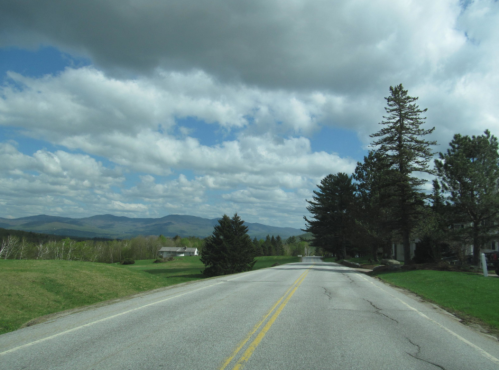 A scenic road stretches into the distance, flanked by trees and mountains under a partly cloudy sky.