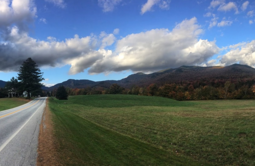 A scenic view of a winding road beside green fields, with mountains and colorful autumn trees under a cloudy sky.