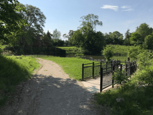 A grassy path leads to a fenced area by a calm river, surrounded by trees and greenery under a clear blue sky.