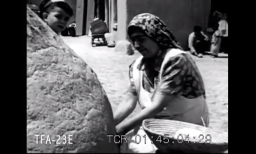 A woman in traditional attire works near a large stone, while a child watches in a historical black-and-white scene.