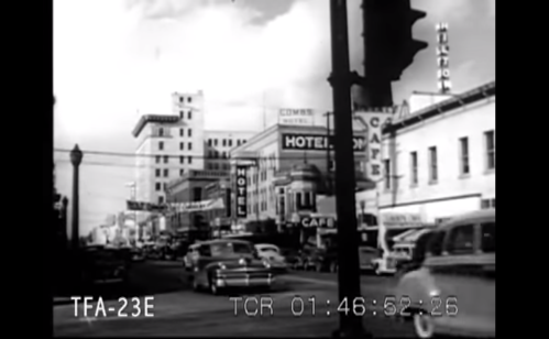 Black and white street scene with cars, buildings, and signs, depicting a busy urban area in the past.