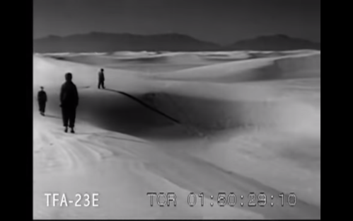 Black and white image of four figures walking across a vast, sandy landscape with rolling dunes.