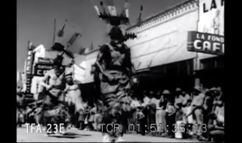 Black and white image of a parade with dancers in traditional costumes and spectators in the background.