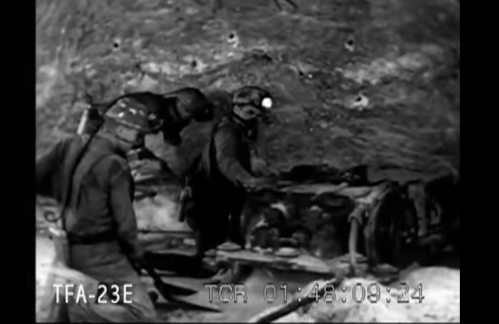 Black and white image of miners working underground, examining machinery in a rocky tunnel.