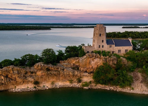 A scenic view of a stone tower on a rocky cliff by a lake, with boats navigating the water under a colorful sky.