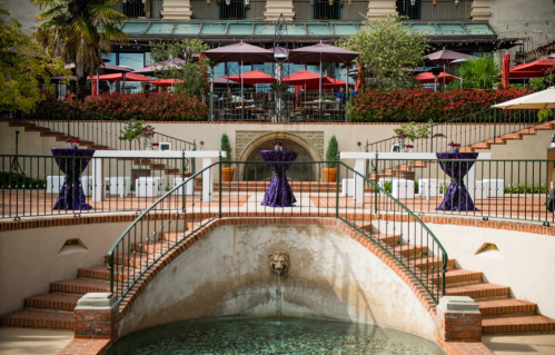 Elegant outdoor venue with purple tablecloths, a fountain, and red umbrellas, surrounded by lush greenery and flowers.