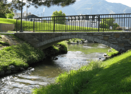 A stone bridge arches over a calm stream, surrounded by green grass and trees, with mountains in the background.