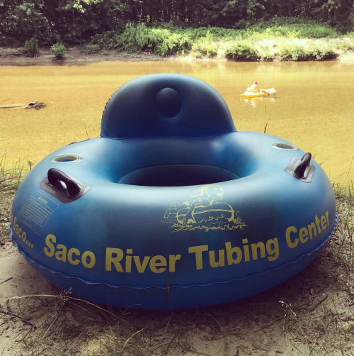 A blue inflatable tube with "Saco River Tubing Center" printed on it, near a calm river with a person kayaking in the background.