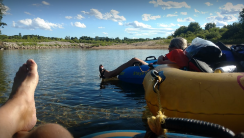 A person relaxes on a tube in a calm river, with another tube nearby and a scenic sky in the background.