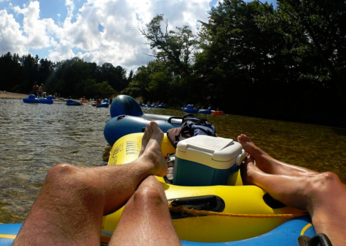 Two pairs of feet resting on a yellow inflatable raft in a river, with other rafts and trees in the background.