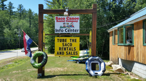 Sign for Saco Canoe Rental Company with info center, featuring tubing rentals and sales, surrounded by greenery.