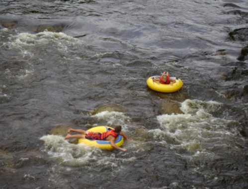 Two people float on yellow inner tubes in a river, surrounded by rocky banks and flowing water.