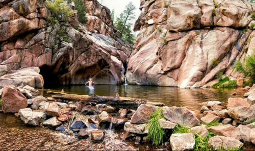 A person jumps into a serene pool surrounded by large rock formations and lush greenery.