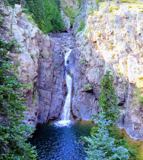 A serene waterfall cascades down rocky cliffs into a clear pool, surrounded by lush greenery and towering trees.