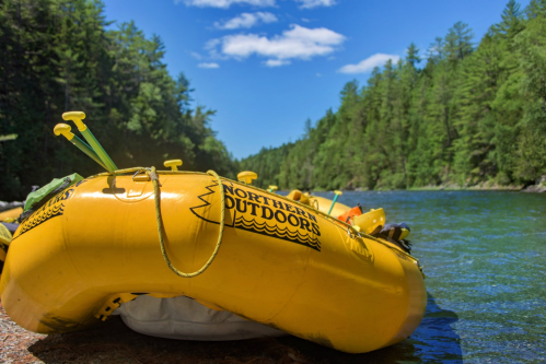 A yellow raft labeled "Northern Outdoors" rests by a river surrounded by lush green trees under a clear blue sky.