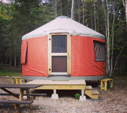A red yurt surrounded by trees, with a wooden deck and picnic table in front.