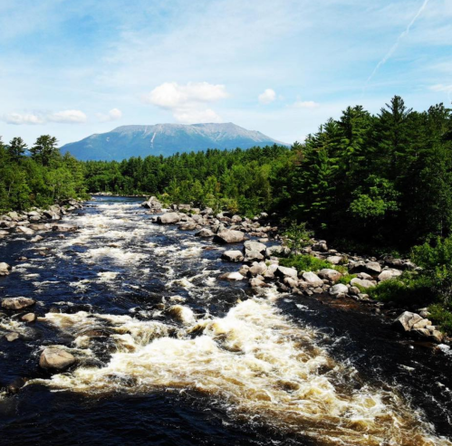 A scenic river flows through a forested landscape, with a mountain in the background under a blue sky.