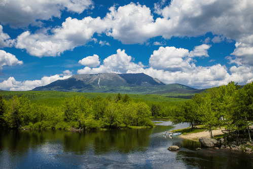A scenic view of a mountain surrounded by lush greenery and a calm river under a blue sky with fluffy clouds.