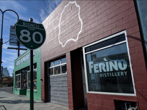 Exterior of Ferino Distillery with a green building nearby and a Business Loop 80 sign in the foreground.