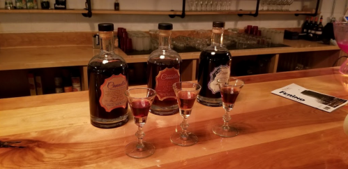 Three bottles of liquor on a wooden bar, with four small glasses filled with dark liquid in front.