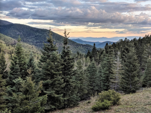A scenic view of lush green mountains under a cloudy sky at sunset, with layers of hills in the background.