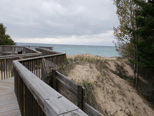 A wooden walkway leads to sandy dunes and a calm lake under a cloudy sky.