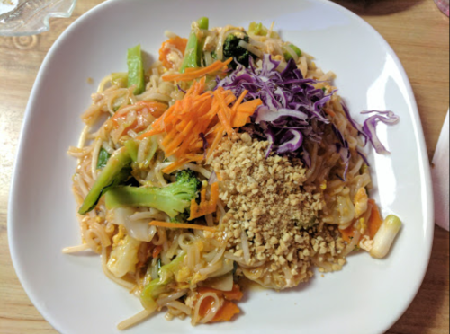A plate of colorful stir-fried noodles topped with vegetables, shredded carrots, purple cabbage, and crushed peanuts.