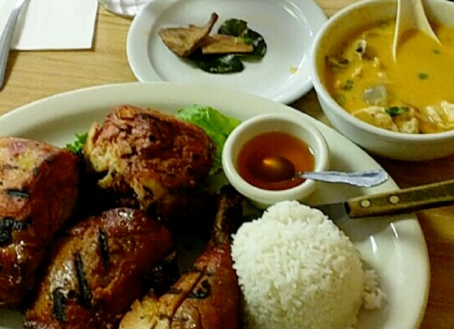 A plate of grilled chicken with rice, a small bowl of soup, and a side of green chilies on a wooden table.