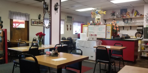 Cozy restaurant interior with wooden tables, black chairs, and colorful decor, featuring a service counter and flowers.