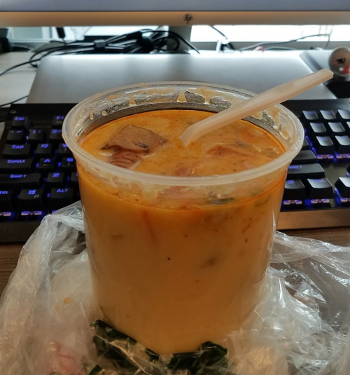 A plastic cup filled with creamy soup, topped with chunks of meat, sitting on a desk with a keyboard in the background.