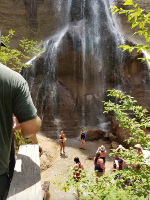People enjoying a waterfall in a lush, green setting, with some wading in the water and others observing from a path.