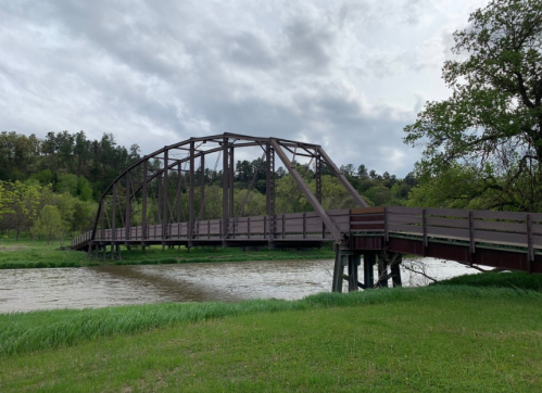A wooden bridge arches over a river, surrounded by green grass and trees under a cloudy sky.