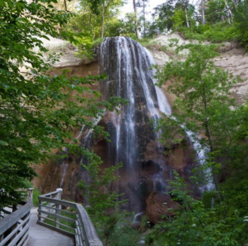 A serene waterfall cascading down a rocky cliff, surrounded by lush greenery and a wooden walkway.